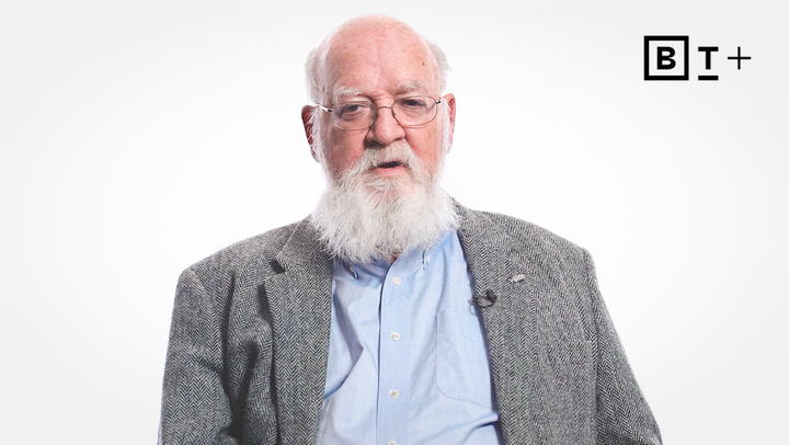 An older man with a white beard and glasses wears a gray jacket and light blue shirt, sitting against a plain white background with a "BT+" logo in the corner.