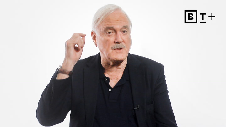 An older man with white hair and a mustache gestures with his right hand while speaking. He is wearing a black jacket and shirt against a plain white background with a "BT+" logo.