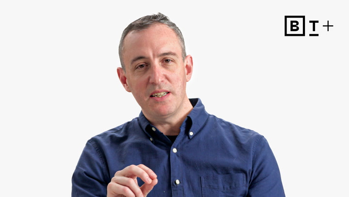 A man in a blue button-up shirt speaks while gesturing with his hand; a "B T +" logo appears in the top right corner on a plain white background.