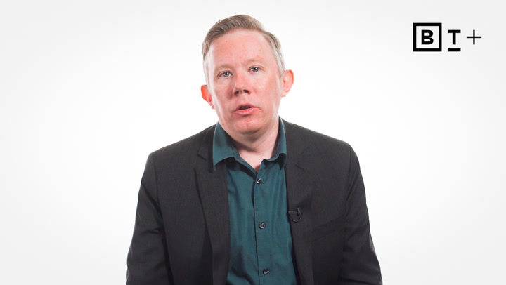 A person in a dark suit and green shirt sits against a plain white background with a B T plus logo in the top right corner.