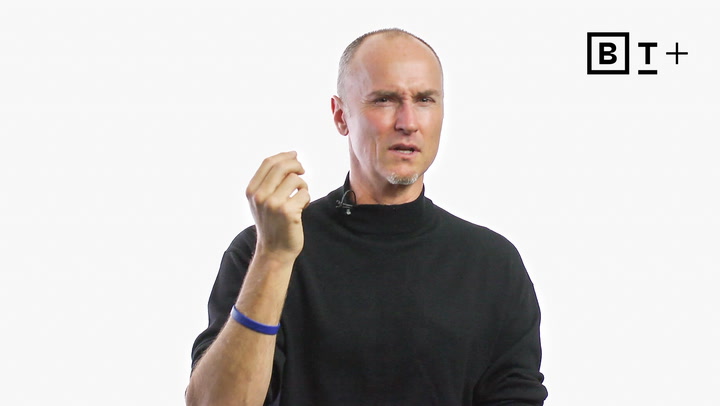 A man in a black turtleneck gestures with his right hand while speaking, with a microphone clipped to his collar and a logo reading "BT+" in the top right corner.