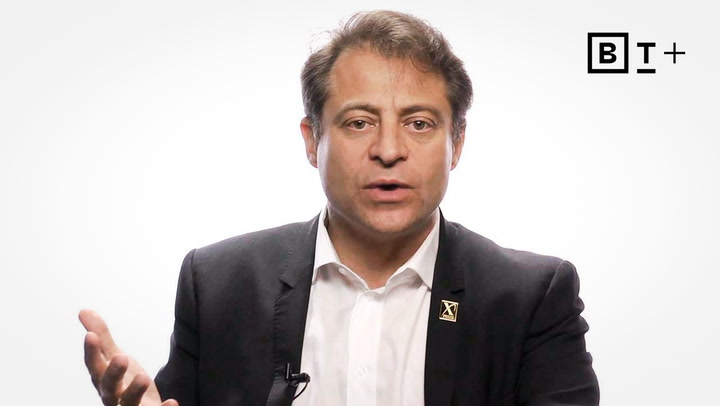 A man in a suit speaks while gesturing with one hand; a "BT+" logo appears in the top right corner against a white background.