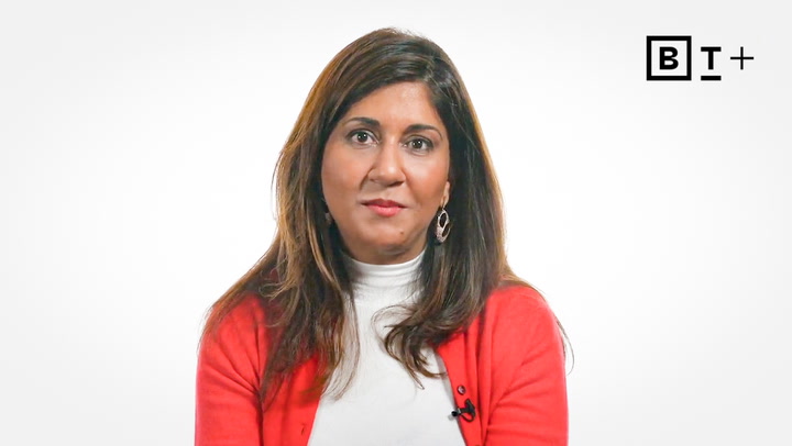 A woman with long brown hair wearing a white turtleneck and red sweater sits in front of a white background with a "B T +" logo in the top right corner.