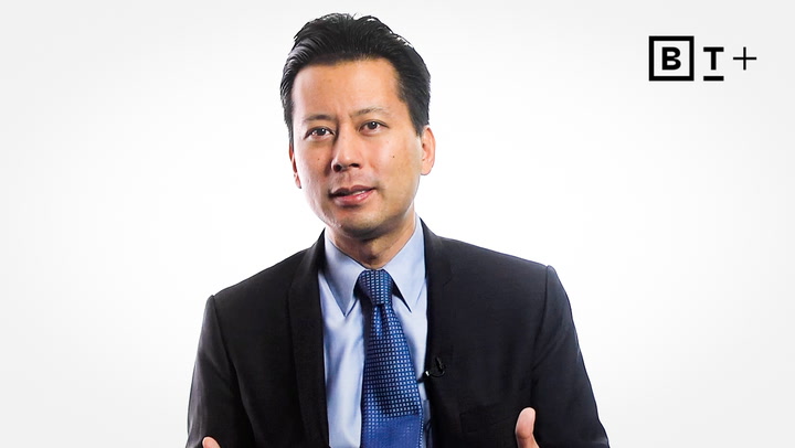 A man in a dark suit and blue tie speaks in front of a white background with a "B T +" logo in the top right corner.