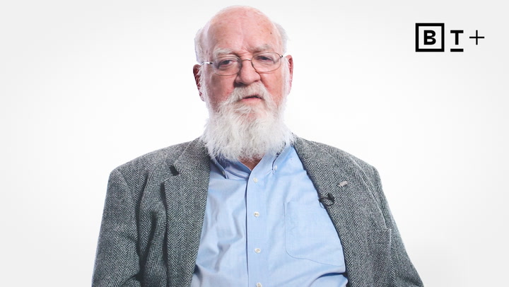 An older man with a white beard and glasses wears a gray jacket and light blue shirt, seated against a plain white background with a "BT+" logo in the corner.