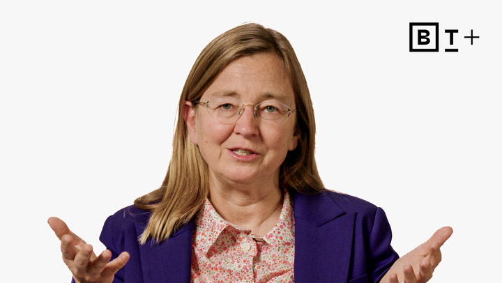 A woman with glasses and long light brown hair, wearing a purple blazer and floral blouse, gestures with her hands in front of a white background with a "B T +" logo.