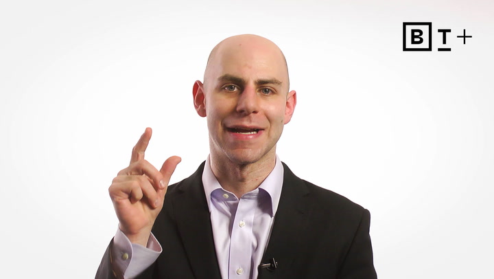 A man in a suit gestures with his right hand while speaking, standing against a plain white background with a "B T+" logo in the top right corner.