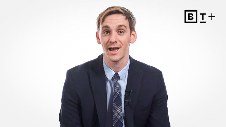 A man in a suit and tie sits in front of a white background, speaking. The letters "B T +" appear in the top right corner.