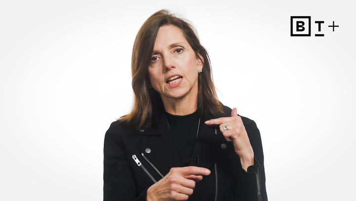 Woman with shoulder-length brown hair wearing a black jacket speaks while gesturing with her hands; BT+ logo appears in the top right corner against a white background.