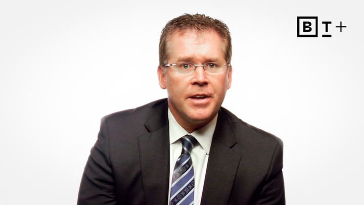 A man in a suit and tie sits against a plain white background with a "BT+" logo in the top right corner.