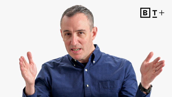 A man wearing a blue button-up shirt gestures with both hands while speaking. A black and white "B T +" logo appears in the top right corner.