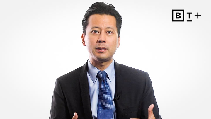 A man in a suit and blue tie speaks in front of a white background with a BT+ logo in the upper right corner.