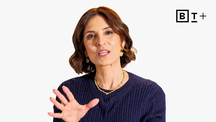 A woman with shoulder-length brown hair wearing a navy sweater and gold jewelry gestures with her hand against a plain white background with a "BT+" logo in the top right corner.