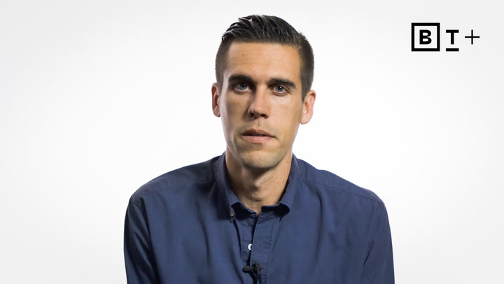A man with short dark hair wearing a blue button-up shirt sits against a plain white background with a "B T +" logo in the top right corner.