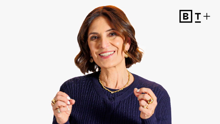 A woman with shoulder-length brown hair, wearing a navy sweater and gold jewelry, gestures while smiling in front of a white background with a "BT+" logo in the top right corner.