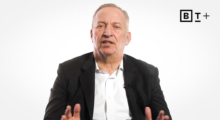 A man in a suit speaks while gesturing with his hands, seated against a plain white background with a "B T+" logo in the top right corner.