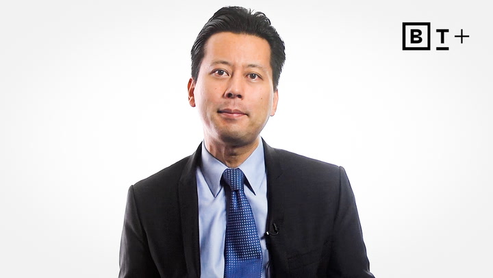 A man in a suit and blue tie sits in front of a white background with a "BT+" logo in the top right corner.