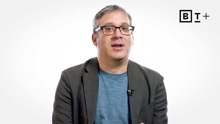 A person wearing glasses, a blue shirt, and a dark blazer speaks in front of a plain white background with a BT+ logo in the top right corner.