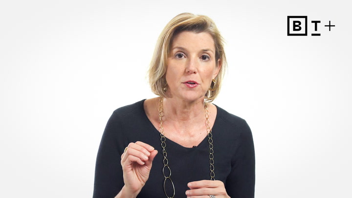 A woman with short blonde hair, wearing a black top and gold necklace, speaks while gesturing with her hand. A "BT+" logo appears in the upper right corner on a plain white background.
