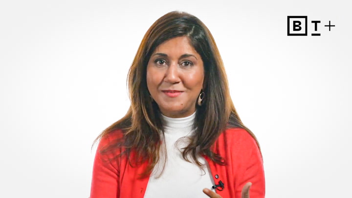 A woman with long brown hair, wearing a white turtleneck and red cardigan, is sitting and smiling in front of a plain white background with a "BT+" logo in the corner.