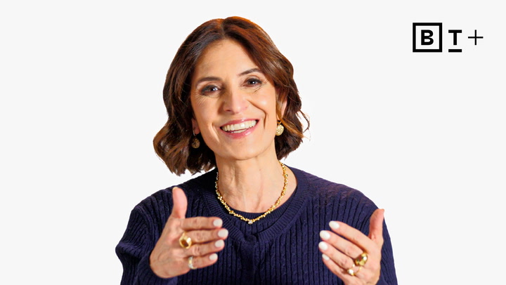 A woman with short brown hair wearing a navy sweater and gold jewelry gestures with her hands and smiles; a "B T +" logo is in the top right corner.