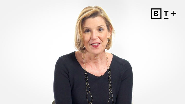 A woman with short blonde hair, wearing a black top and gold necklace, sits in front of a plain white background with a "BT+" logo in the upper right corner.