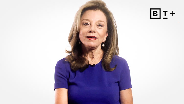 A woman with long brown hair, wearing a blue short-sleeve top, sits in front of a plain white background with a black "BT+" logo in the upper right corner.