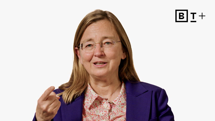 A woman with glasses and long brown hair wearing a purple blazer and floral shirt gestures with her hand, set against a plain white background with a "B T +" logo in the corner.