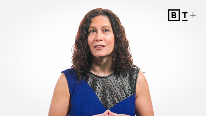 A woman with curly brown hair in a blue dress stands against a white background with a "BT+" logo in the corner.