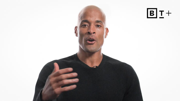 A man in a black sweater speaks while gesturing with his hand. A "B T +" logo appears in the top right corner on a plain white background.