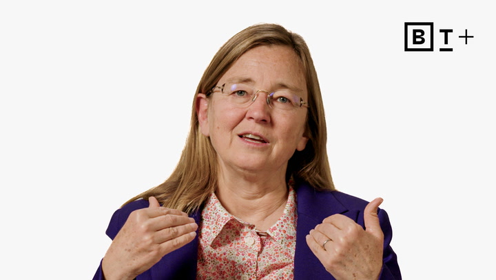A woman with long light brown hair, glasses, and a purple blazer gestures with both hands while speaking, against a plain white background with the B T+ logo in the top right corner.