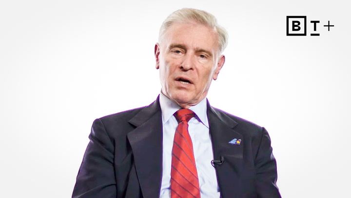 An older man in a suit and red tie sits against a plain white background. A "B T +" logo is displayed in the top right corner.