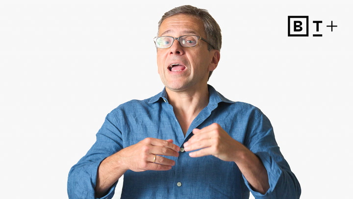 A man wearing glasses and a blue shirt gestures with his hands while speaking; a black and white "B T +" logo is in the top right corner.