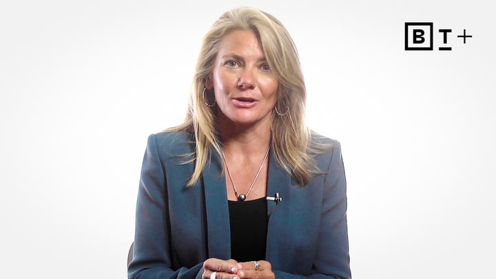 A woman with blonde hair in a blue blazer sits and speaks in front of a plain white background with a "BT+" logo in the corner.