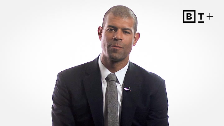 A man in a dark suit and tie sits against a plain white background. The BT+ logo is visible in the upper right corner.