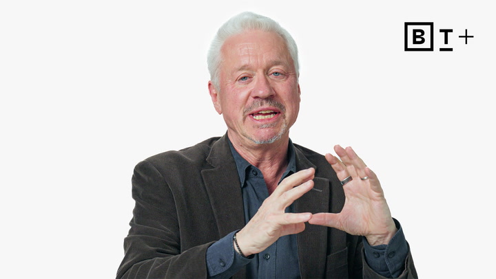 An older man with white hair and a beard speaks while gesturing with his hands. He is wearing a dark jacket and shirt. A "B T +" logo appears in the top right corner.