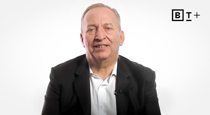 A man in a dark suit and white shirt sits in front of a plain white background with a "B T +" logo in the top right corner.