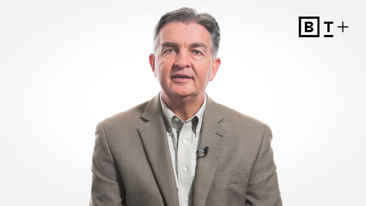 A middle-aged man in a tan blazer and light shirt sits facing the camera against a plain white background with a "BT+" logo in the top right corner.