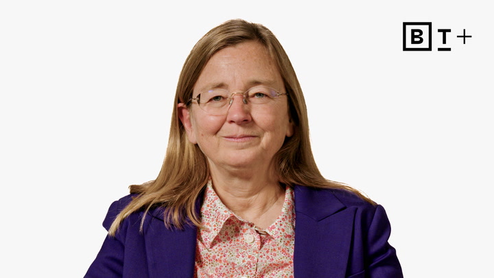 A woman with long light brown hair and glasses, wearing a purple blazer and floral shirt, sits in front of a white background with the B+ logo in the corner.
