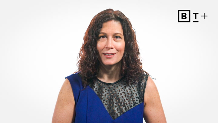 A woman with curly brown hair wearing a blue sleeveless top is standing in front of a white background with a "B T +" logo in the top right corner.