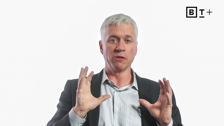 A man in a suit gestures with both hands while speaking in front of a plain white background with a "B T +" logo in the top right corner.