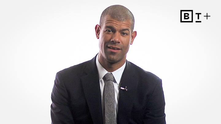 A man in a dark suit and tie sits against a plain white background. A "B T +" logo appears in the top right corner of the image.