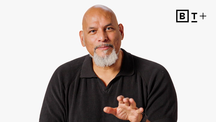 A bald man with a gray goatee wearing a black shirt gestures with his hand, with a "B T+" logo in the top right corner against a white background.