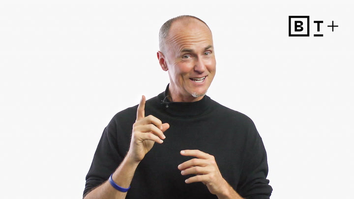 A man in a black turtleneck gestures with one hand raised, smiling. A logo with the letters "B T +" appears in the top right corner on a white background.