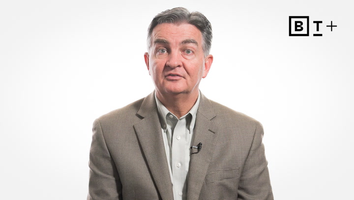 A man in a brown blazer and light shirt sits facing the camera against a plain white background, with a "B T +" logo in the top right corner.