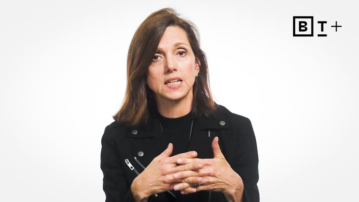 A woman with shoulder-length brown hair, wearing a black jacket, speaks while gesturing with her hands. A "BT+" logo is visible in the top right corner against a white background.