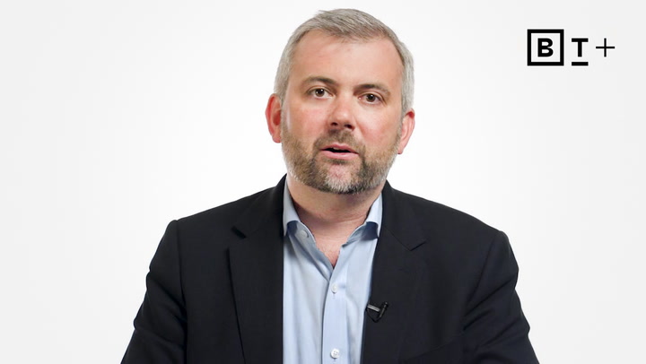 A man in a suit and open-collared shirt speaks in front of a plain white background with a "B T +" logo in the top right corner.