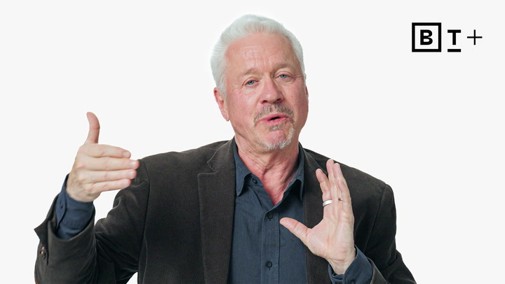 An older man with gray hair and a goatee gestures with his hands while speaking, wearing a dark blazer and shirt. A "B T +" logo appears in the top right corner.