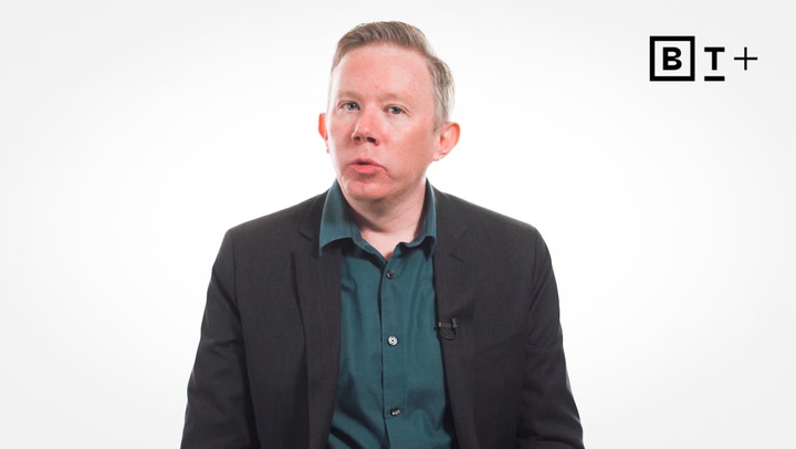 A person wearing a dark blazer and teal shirt sits in front of a plain white background with a "BT+" logo in the corner.