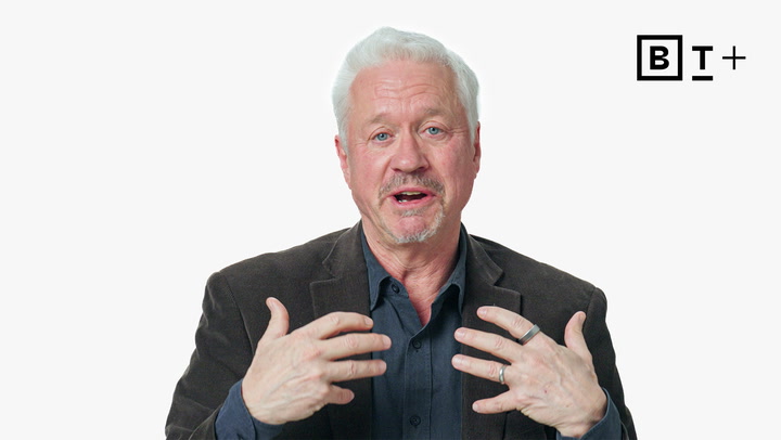 An older man with white hair and a beard, wearing a dark blazer and shirt, gestures with both hands while speaking. A "B T +" logo appears in the top right corner on a plain white background.